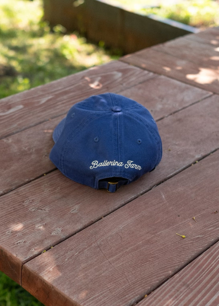 Blue baseball cap with adjustable strap and embroidered Ballerina Farm logo rests on a weathered wooden outdoor table.